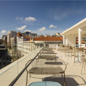 Terraza moderna con sillas, mesas y vista urbana bajo un cielo azul.